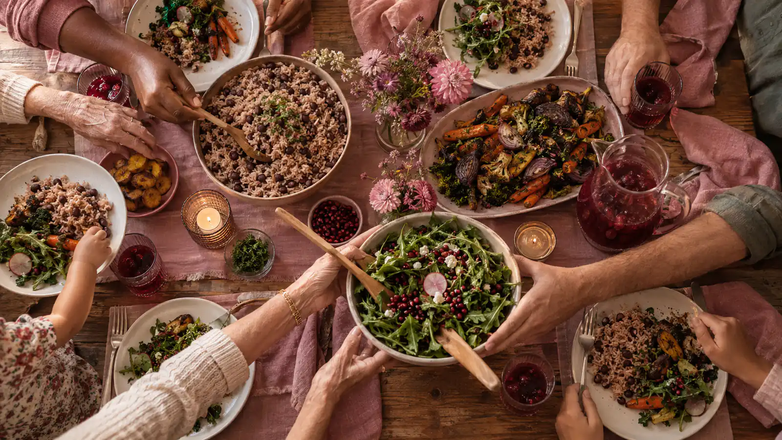 Hands of all ages passing bowls of rice and peas, roasted vegetables and salad around a family dinner table, seen from above